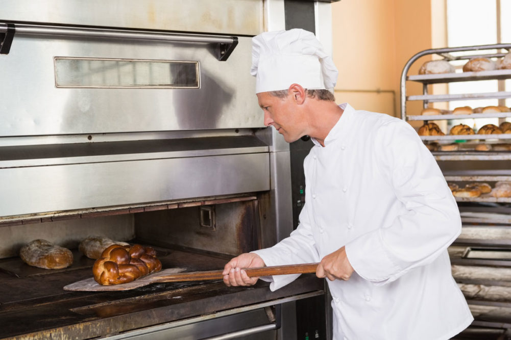 Baker in white coat removing bread from oven. 